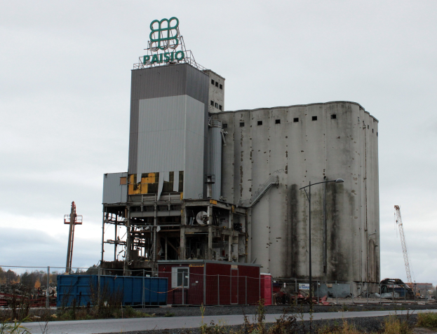 Großer Zementbau neben einer Straße, umgeben von Pflanzen, einem Zaun, Pfählen, Lampen und anderen Gegenständen, mit Bäumen und einem klaren blauen Himmel im Hintergrund.