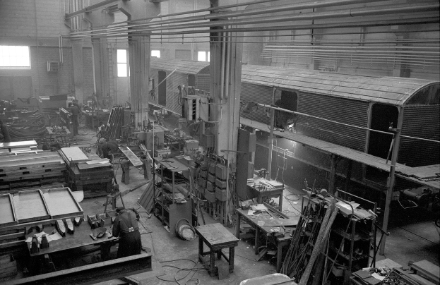 Schwarzes und weißes Foto eines Zuges in einer Fabrik mit Arbeitern, verstreuter Ausrüstung und sichtbaren Wänden und Fenstern.