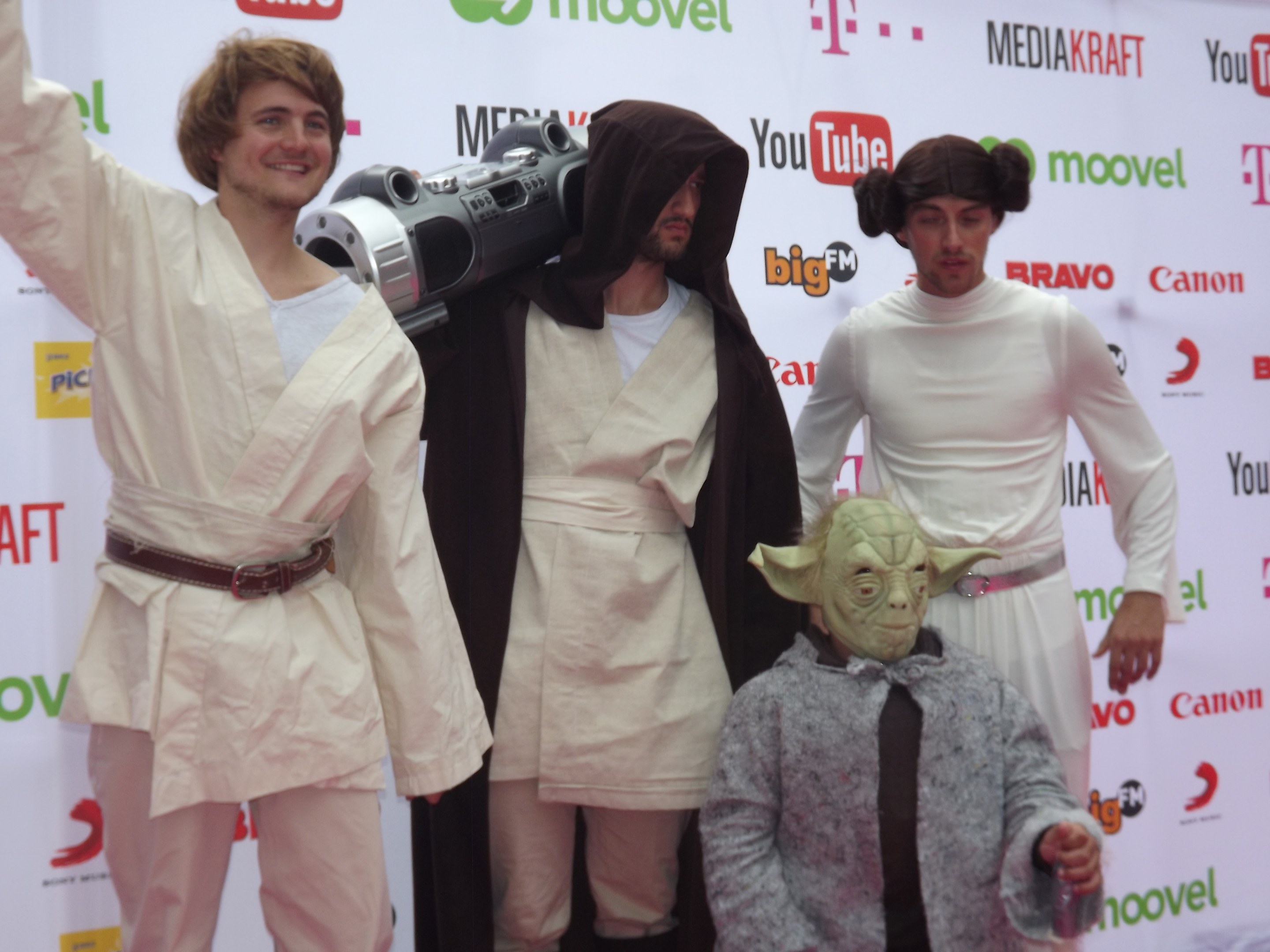 Eine Gruppe von Menschen in Star-Wars-Kostümen, die auf einem roten Teppich posen, wobei einer ein Objekt und ein Banner mit Text und Logos im Hintergrund hält.