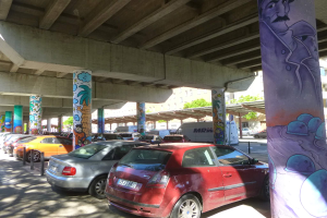 Ein Parkplatz voller parkender Autos unter einer Brücke, mit Graffiti an den Säulen und Gebäuden und Bäumen im Hintergrund, mit einem Van.