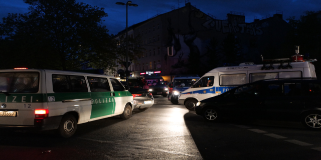 Eine Gruppe von Polizeiwagen, darunter ein Van, ein Auto und ein Rad, geparkt am Straßenrand bei Nacht, mit Gebäuden, Bäumen, Laternen und einem sternenklaren Himmel im Hintergrund.