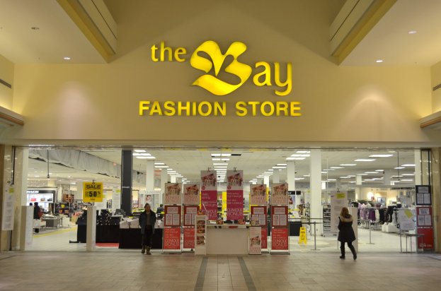 Vorbau eines Bay Fashion Retail Shops in einem Einkaufszentrum mit vorbeigehenden Einkäufern, Texttafeln an den Wänden, tragenden Säulen, Deckenbeleuchtung und zusätzlichen Einkaufszentralelementen im Hintergrund.