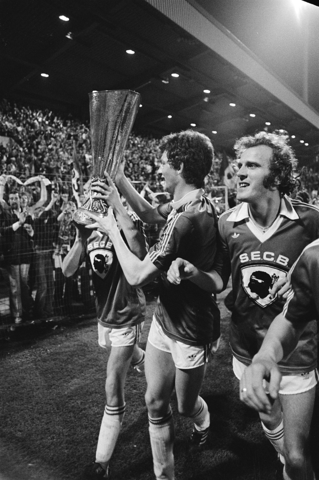 Schwarzes und weißes Foto von Fußballspielern, die mit einem Pokal auf einem Feld feiern, umgeben von einer jubelnden Menge hinter einem Zaun unter Stadionbeleuchtung.