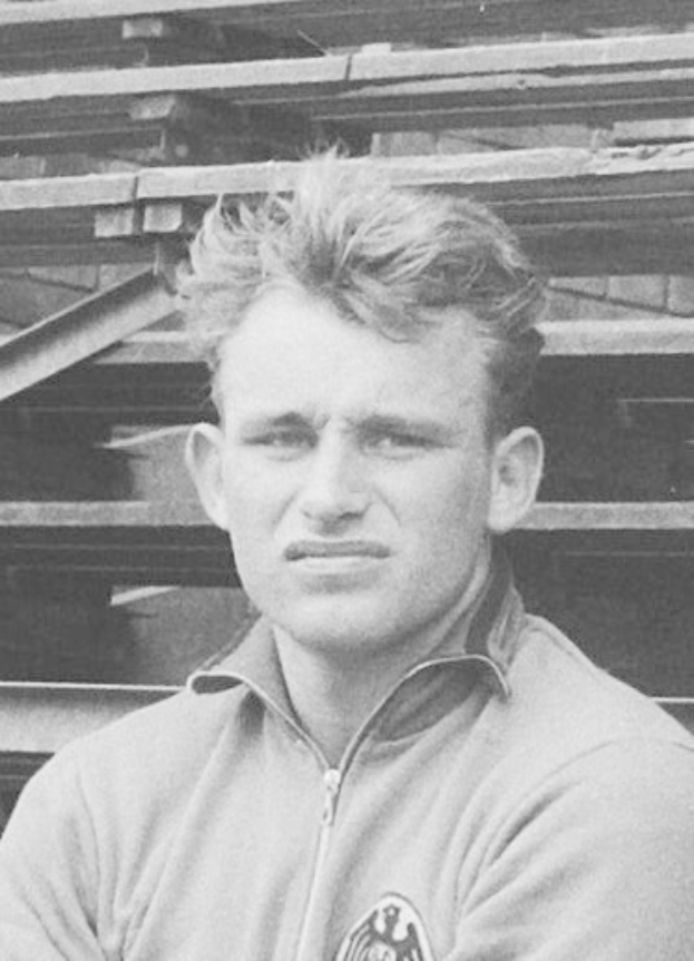 Schwarzes und weißes Foto eines Mannes in einer Jacke, der vor gestapelten Holzplanken steht.