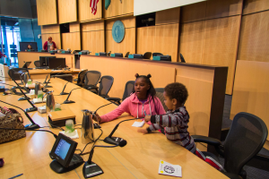 Zwei Kinder sitzen an einem langen Tisch mit Mikrofonen, Flaschen, Taschen und Papieren und führen ein Gespräch bei einer Schulkonferenz, mit Menschen und Fahnen im Hintergrund.