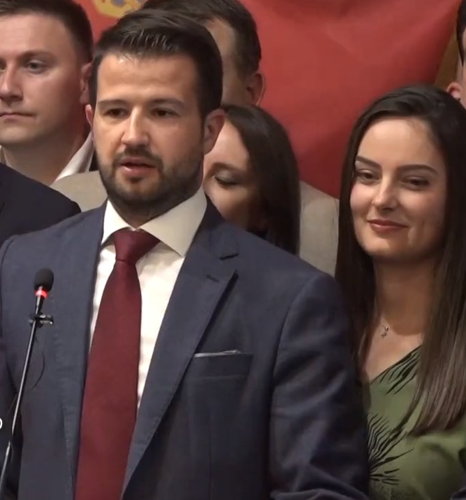 Ein Mann im Anzug spricht in ein Mikrofon an einem Rednerpult, mit einem Banner dahinter und einer Menge davor, wahrscheinlich zu einem Regierungssozialmedienverbot.