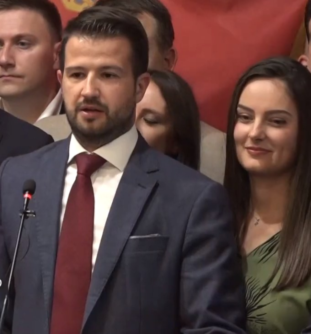 Ein Mann im Anzug spricht in ein Mikrofon an einem Rednerpult, mit einem Banner dahinter und einer Menge davor, wahrscheinlich zu einem Regierungssozialmedienverbot.