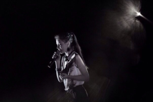 Eine Frau in einem schwarzen und weißen Outfit singt leidenschaftlich auf einer von Scheinwerfern beleuchteten Bühne, die Augen geschlossen, ein Mikrofon haltend.