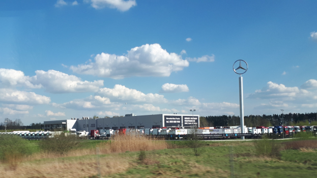 Ein Mercedes-Benz-Werk in Deutschland mit Fahrzeugen auf der Straße, Grün, Gebäuden und einem Logo-Mast unter einem bewölktem Himmel.