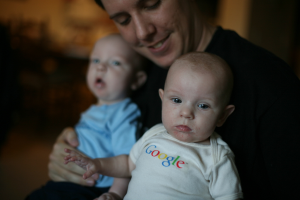 Eine Person, die zwei Babys hält, mit lesbarem Text auf dem Kleid des vorderen Babys und einem unscharfen Hintergrund.