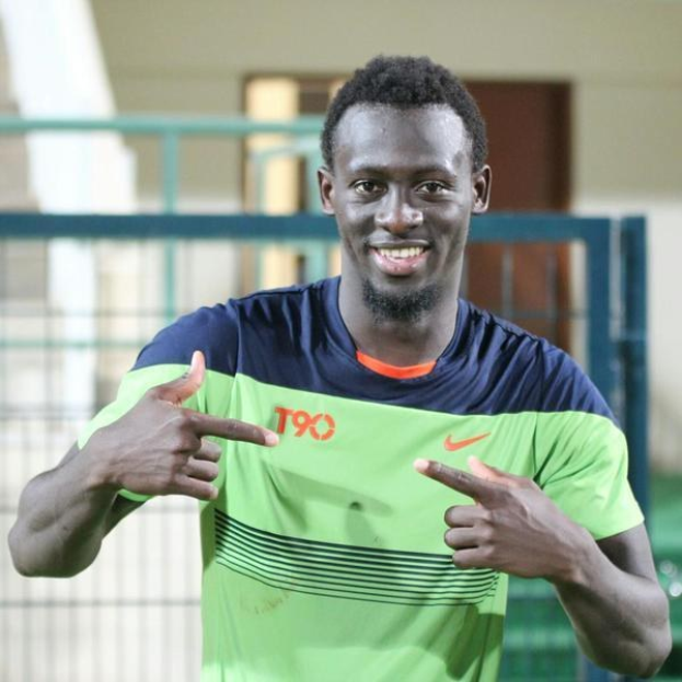 Ein Mann in einem grünen und blauen Hemd, der in die Kamera lächelt und auf sie zeigt, mit einem unscharfen Geländer, einer Wand und einer Tür im Hintergrund.