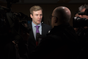 Ein Mann im Anzug und mit Krawatte, der eine Brille trägt, spricht mit Reportern, während zwei Kameramitarbeiter rechts und links von ihm stehen und ein Mikrofon vor ihm platziert ist.