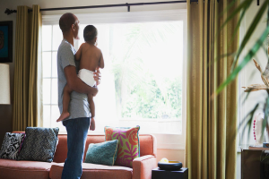 Ein Mann trägt ein Kind vor einem Fenster mit Vorhängen, einer Couch und mehreren Kissen auf der Couch.