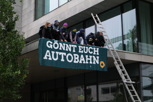 Eine Gruppe von Menschen auf einem Gebäude, die eine Fahne halten, mit einem Baum links und Glasfenstern im Hintergrund, die scheinbar protestieren und die deutsche Regierung unterstützen.