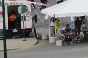 Ein Krankenwagen steht links von einer Straße, während rechts ein Zelt eine Gruppe von Menschen schützt, mit einem Tisch und einem Stuhl in der Nähe.