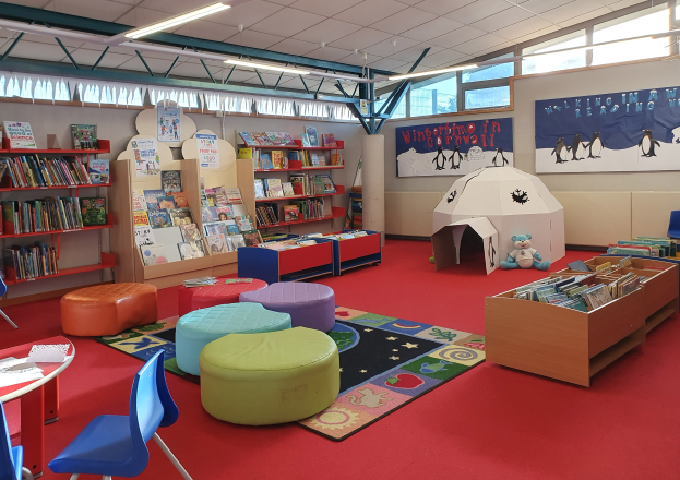 Eine hell erleuchtete Kinderbibliothek mit Bücherregalen, Tischen, Stühlen, Teppichen, Wandtafeln und großen Fenstern.