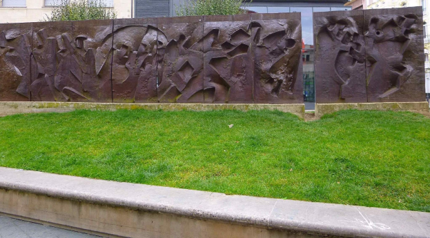 Große Metallskulptur der Berliner Mauer auf einer grünen Wiese umgeben von Bäumen und Gebäuden mit Fenstern im Hintergrund.