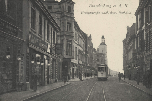 Ein Schwarz-Weiß-Foto einer Stadtstraße in Brandenburg, Deutschland, das eine Tram auf Schienen, Menschen auf dem Bürgersteig, Gebäude mit Fenstern, Schilder und Straßenlaternen unter einem bewölkten Himmel zeigt.