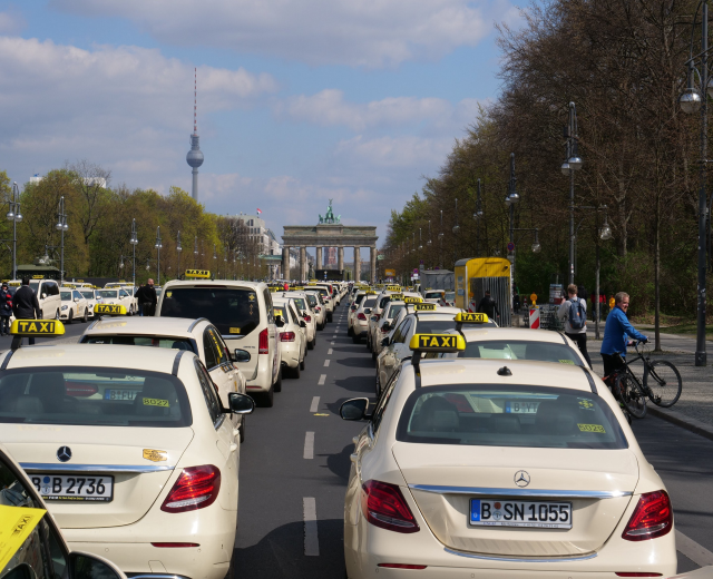 Eine lange Reihe von Taxis steht entlang einer belebten Straße in Berlin, Deutschland, mit Fahrzeugen, Radfahrern und Fußgängern, flankiert von Laternen und Bäumen und Gebäuden, einem Bogen und einem Turm im Hintergrund unter einem bewölkten Himmel.