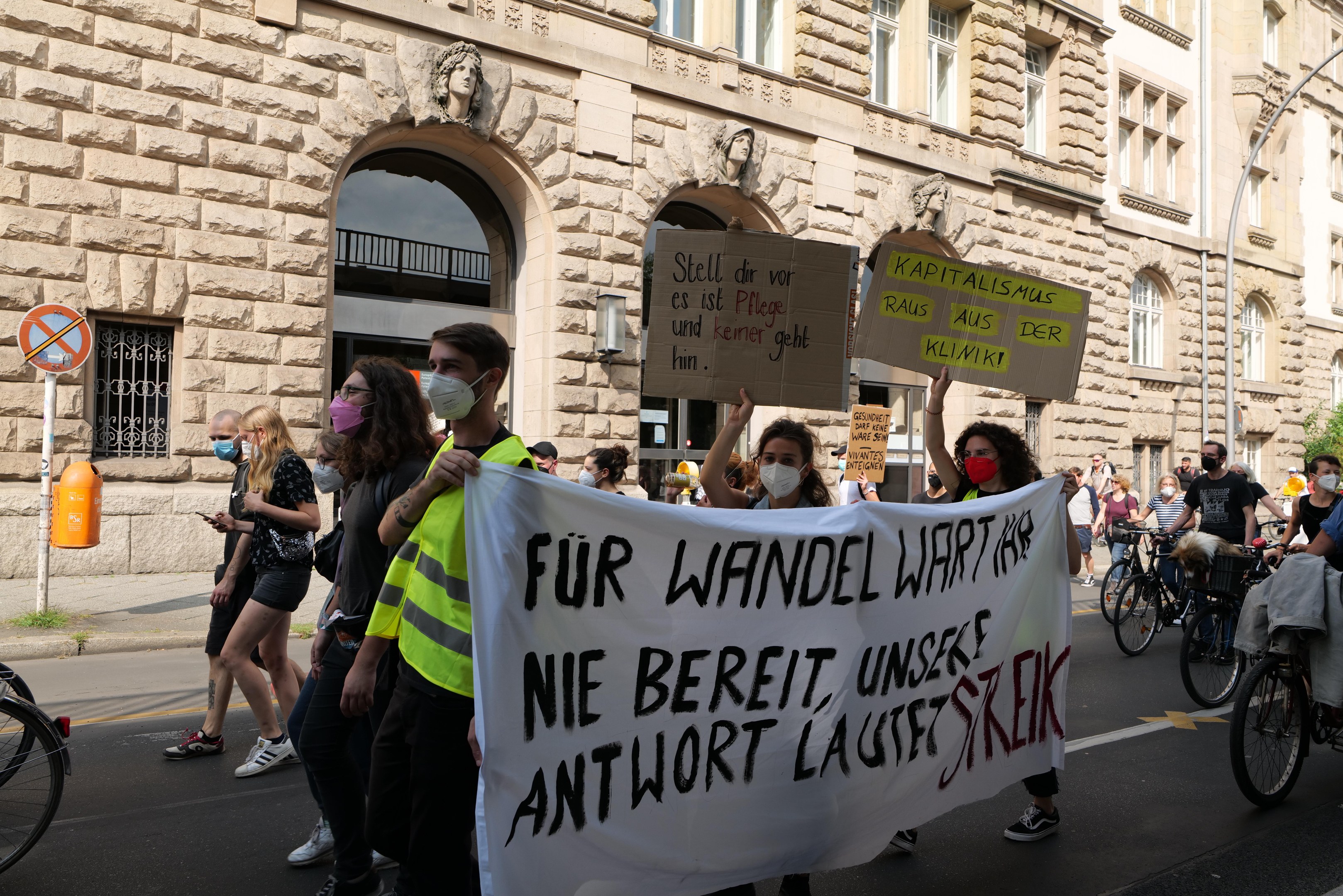 Eine Gruppe von Menschen marschiert bei einer Demonstration auf einer Stadtstraße, einige halten Schilder und andere fahren Fahrräder, mit einem historischen Gebäude im Hintergrund, das gewölbte Fenster, Säulen und Skulpturen aufweist.