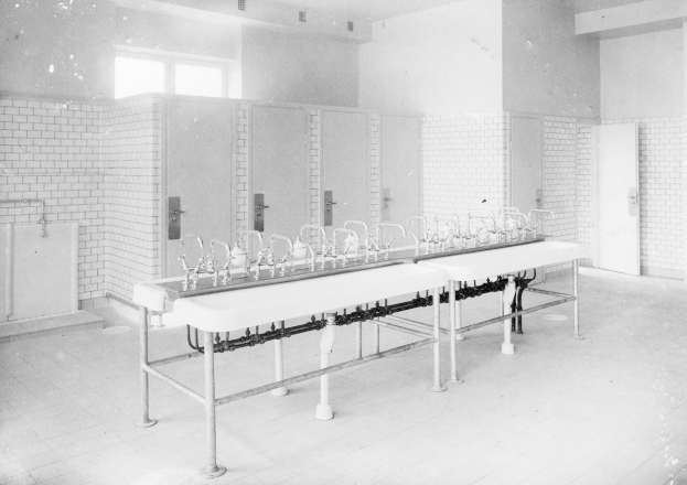 Schwarz-weiß-Foto einer öffentlichen Toilette mit einer Reihe von Waschbecken, Wasserhähnen, Wänden, Türen, Fenstern und einem gefliesten Boden, mit verschiedenen Gegenständen, die herumliegen.