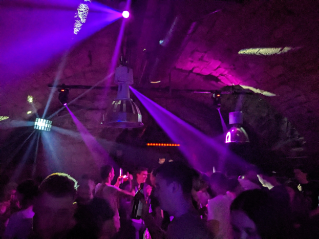 Eine Gruppe von Menschen, die in einer Diskothek tanzen, unter lila und blauen Deckenlichtern, einige halten Flaschen.