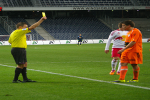 Ein Schiedsrichter zeigt einem Fußballspieler auf dem Feld eine gelbe Karte, umgeben von einer Gruppe von Menschen, mit einem Torpfosten, einem Zaun, einer Tafel, Metallrahmen, einem Anzeigebildschirm, Stühlen und einem Dach mit Deckenleuchten im Hintergrund.