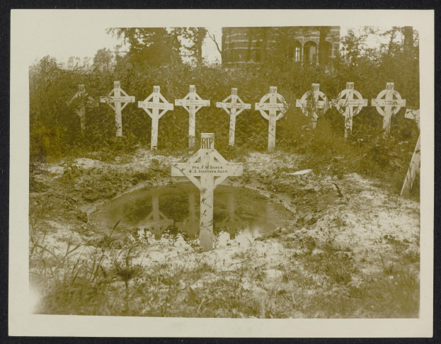 Schwarz-weiß-Foto eines Friedhofs mit zahlreichen Kreuzen im Vordergrund, umgeben von Bäumen und einem Gebäude im Hintergrund, mit einem Gewässer in der Mitte.