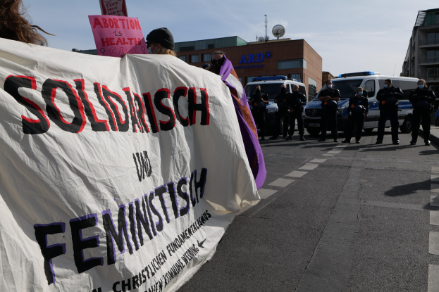 Gruppe von Menschen marschiert auf einer Straße mit einem Banner 'Solidarität und Feminismus' vorbei an parkenden Autos und Gebäuden im Hintergrund unter einem klaren blauen Himmel.