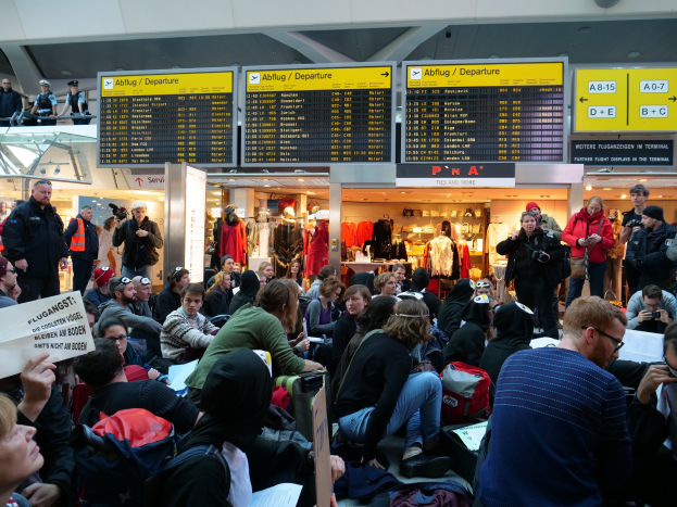 Eine große Gruppe von Menschen in einem Flughafen, einige sitzen mit Taschen und Papieren, andere stehen, mit Hintergrundtafeln, Schaufensterpuppen in Kleidern und Deckenleuchten, was auf eine Demonstration hindeutet.