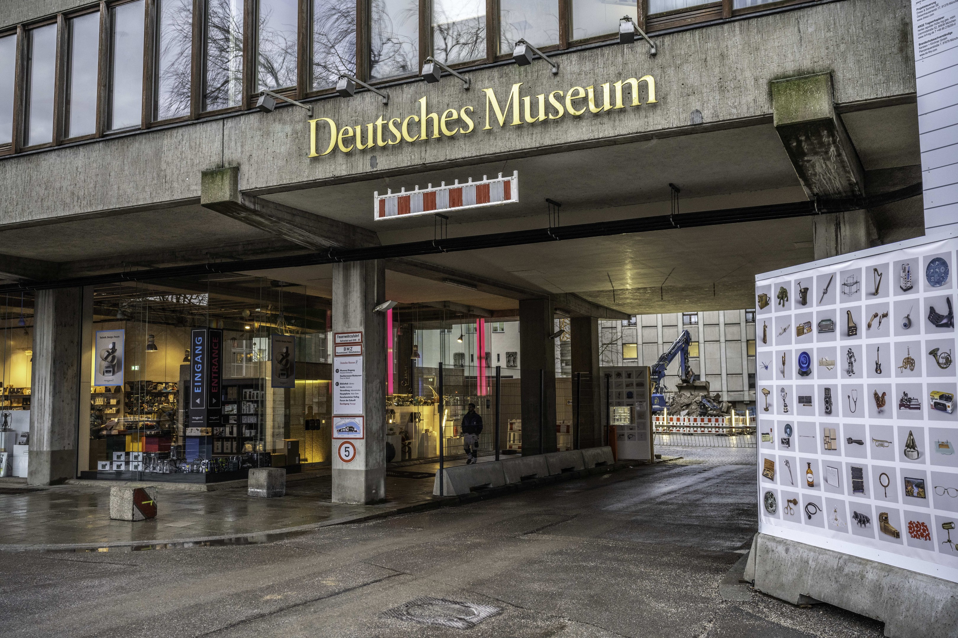 Außenansicht des Deutschen Museums in Berlin, das ein großes Gebäude mit Glasfenstern, Säulen und einer Namensplakette zeigt, zusammen mit einer Informationsanzeigetafel, sowie anderen Gebäuden, Bäumen und dem Himmel im Hintergrund.