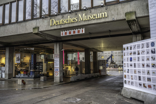 Außenansicht des Deutschen Museums in Berlin, das ein großes Gebäude mit Glasfenstern, Säulen und einer Namensplakette zeigt, zusammen mit einer Informationsanzeigetafel, sowie anderen Gebäuden, Bäumen und dem Himmel im Hintergrund.