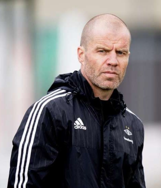 Ein kahlköpfiger Trainer in einer blauen Jacke mit weißen Streifen steht auf einem Fußballfeld mit verschränkten Händen und einem ernsten Gesichtsausdruck.