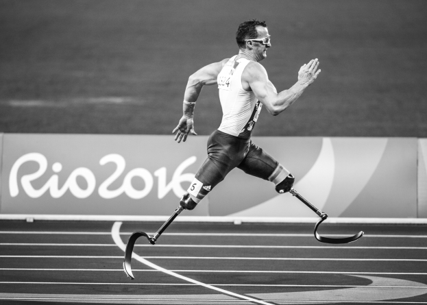 Schwarz-weißes Foto eines Mannes mit einem Prothesenbein, der auf einer Laufbahn läuft, mit einer Tafel im Hintergrund, auf der "Rio 2016 Paralympic Games" steht.
