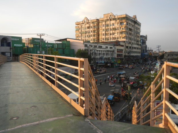 Eine Fußgängerbrücke mit Geländern überspannt eine vielbefahrene Straße voller Fahrzeuge und Fußgänger, mit Gebäuden, Bäumen, Strommasten und einem bewölkten Himmel im Hintergrund.
