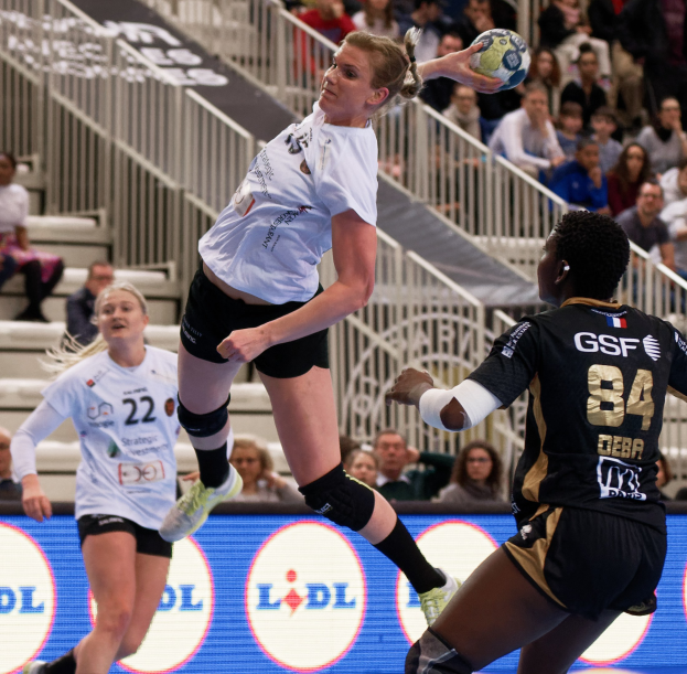 Eine Gruppe von Frauen, die auf einem Platz Handball spielen, mit einem Ball in der Luft, Zuschauern im Hintergrund und einer Tafel mit der Aufschrift "Handball-Weltmeisterschaft 2019 - Tag 2".