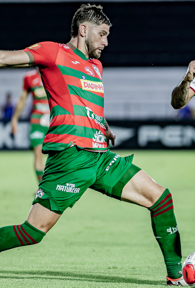 Ein Mann in roter und grüner Uniform kickt einen Fußball auf einem Rasenfeld, mit einer weiteren Person im Hintergrund und einer Tafel mit der Aufschrift "Santos vs Vitoria Guimaraes".