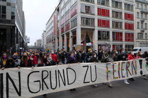 Eine Gruppe von Menschen mit Masken und einem Banner mit der Aufschrift 'In Grund zu Feiern' steht vor Gebäuden, Laternenmasten und Fahrzeugen, was auf eine Demonstration in Berlin, Deutschland, hindeutet.