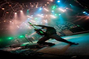 Ein musikalisches Konzert mit drei Personen auf der Bühne: zwei spielen rechts Gitarren mit Mikrofonen davor, ein Hintergrund mit "MOLLEY" geschrieben und zwei Schlangenlogos sowie Scheinwerfer darüber.