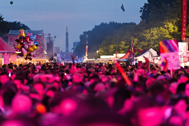 Eine große Menschenmenge auf einem nächtlichen Musikfestival, beleuchtet von bunten Lichtern, mit Zelten, Fahnen und Pfählen im Vordergrund und Bäumen, einem Turm und einem klaren Himmel im Hintergrund.