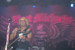 Eine Musik-Konzertszene mit Scheinwerfern oben, einem Banner hinten, Drogen in der Mitte unten, einer sitzenden Person in der rechten unteren Ecke und einem stehenden Gitarristen mit Mikrofon vorne.