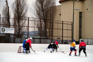 Menschen beim Eishockey auf einem Eisring mit Gebäuden, Bäumen, einer Straßenlaterne, einem Namensschild und Zäunen im Hintergrund unter einem Himmel.