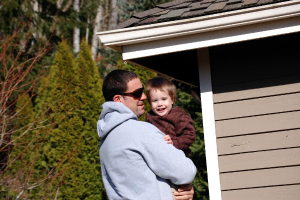 Ein Mann in einer Jacke und Schutzbrille trägt ein Kind, lachend, mit einer Hütte und Bäumen im Hintergrund.