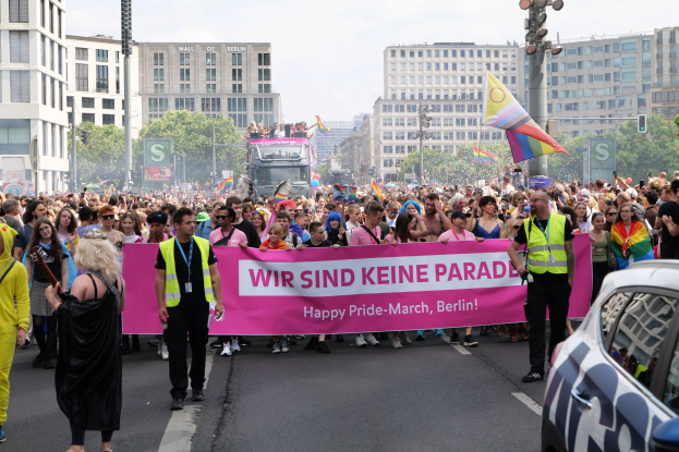 Eine große Gruppe von Menschen marschiert eine Straße entlang und trägt ein Banner mit der Aufschrift "Happy Pride March Berlin", mit einem geparkten Auto auf der rechten Seite, Gebäuden, Bäumen und einem klaren blauen Himmel im Hintergrund.