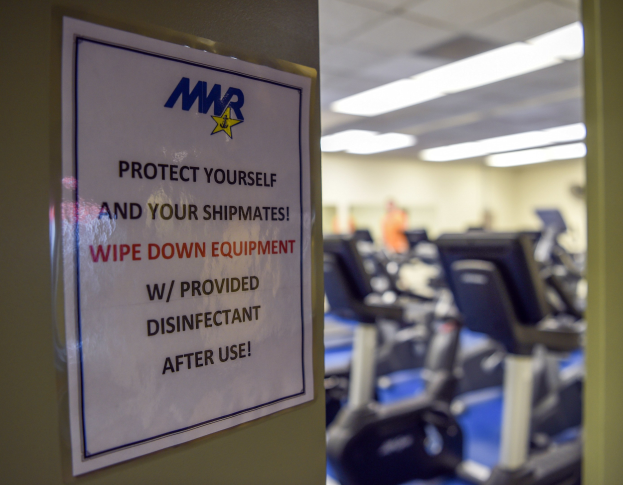 Zeichen an einer Turnhallentür mit der Aufschrift 'Schützen Sie sich und Ihre Schiffskameraden' mit Laufbändern und Deckenlampen im Hintergrund.