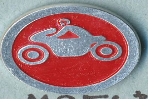 Ein rundes, rotes und silbernes Schild mit einer Person auf einem Motorrad, die einen Helm trägt, mit Text, der wahrscheinlich den Namen des Motorrads angibt.
