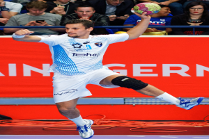 Ein Mann in der Luft mit einem Handball, im Hintergrund eine Gruppe von Menschen und ein Banner mit Text.