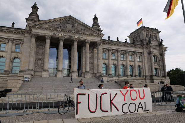 Protestierende halten ein "Fuck You Coal"-Schild vor dem Reichstagsgebäude in Berlin, Deutschland, mit Bäumen, einer Fahnenstange und einem bewölkten Himmel im Hintergrund.