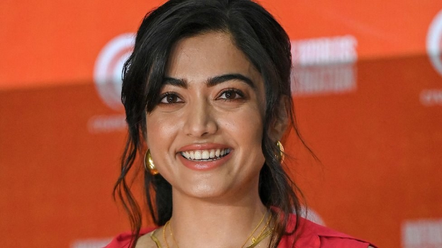 Eine Frau in einem leuchtend roten Kleid mit einem eleganten Dutt und dezentem Make-up steht vor der Kamera und lächelt, trägt schwarze High Heels und hat ein hellgelbes Banner im Hintergrund mit der Aufschrift "Sonam Kapoors beste Schönheits Looks vom Cannes Film Festival."