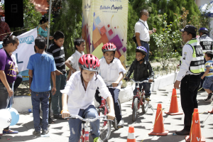 Kinder fahren mit Helmen auf Fahrrädern eine Straße entlang, einige mit Verkehrskegeln, andere in der Nähe stehend, mit einer Fahne, Bäumen und Gebäuden im Hintergrund.